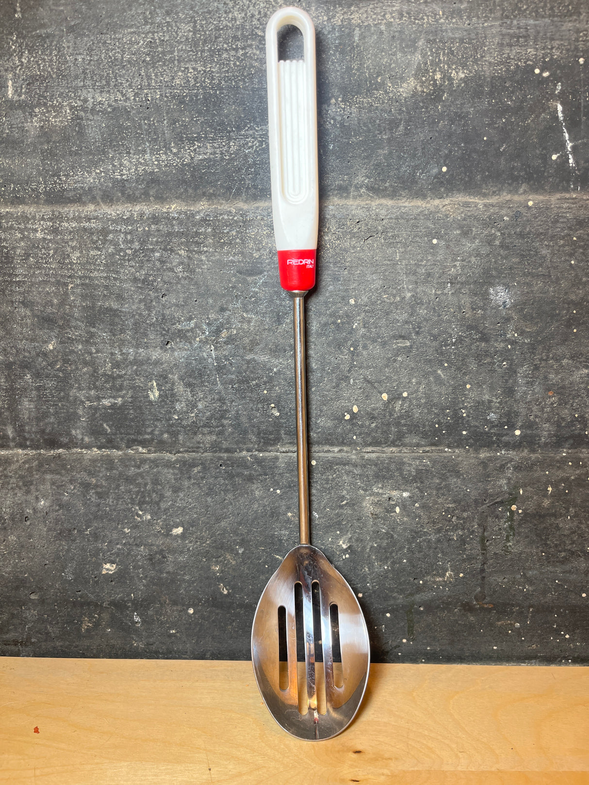 Pedrini stainless steel slotted serving spoon with a long handle, featuring a white plastic grip, red collar, and slender metal shaft for effective draining.