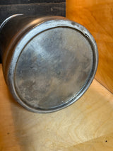 Underside of a vintage John Wood Young’s Dairy stainless steel milk can, showcasing its worn, scratched base with an outward rolled edge, resting on a wooden shelf.
