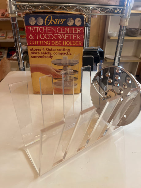 Oster Kitchencenter & Foodcrafter cutting disc holder, clear acrylic, holds four metal discs, includes a stainless-steel blade and original packaging in the background.