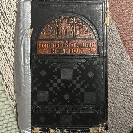 The Household Searchlight Recipe Book displayed on a rug, featuring a black and silver cover, offers tested homemaking recipes from The Household Magazine.