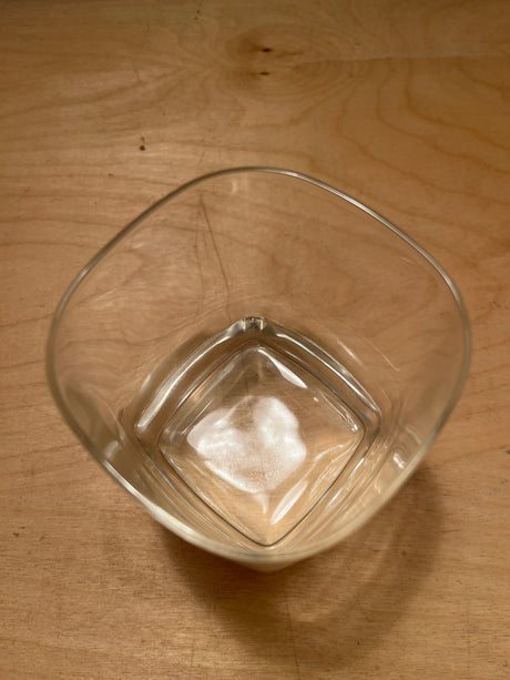 Square heavy bottom tumbler drinking glass on a wooden surface, showcasing its clear, square design and robust base, with light reflections hinting at subtle elegance.
