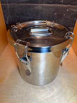 Stainless steel food storage container with clasps, handle, and silicone seal, featuring a cylindrical design with a snap-down locking lid on a wooden shelf.