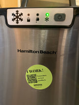 Hamilton Beach portable countertop ice maker with control panel and green snowflake indicator lit, featuring a “Spoons Kitchen Exchange” sticker stating “I WORK!” with a QR code.