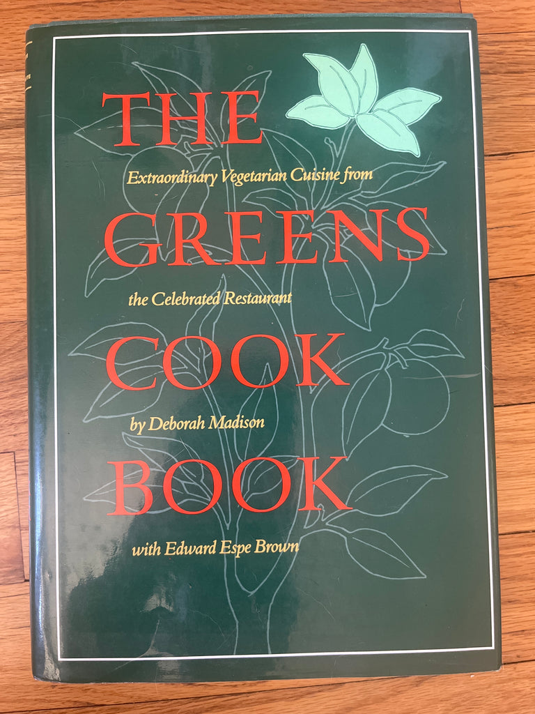 The Greens Cookbook on a wooden surface. The cover features bold red lettering and an illustration of a plant.