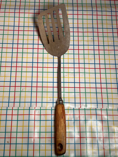Flat thin steel slotted spatula with wood handle - vintage, ideal for flipping pancakes and grilling, shown on a multi-colored grid-patterned surface.