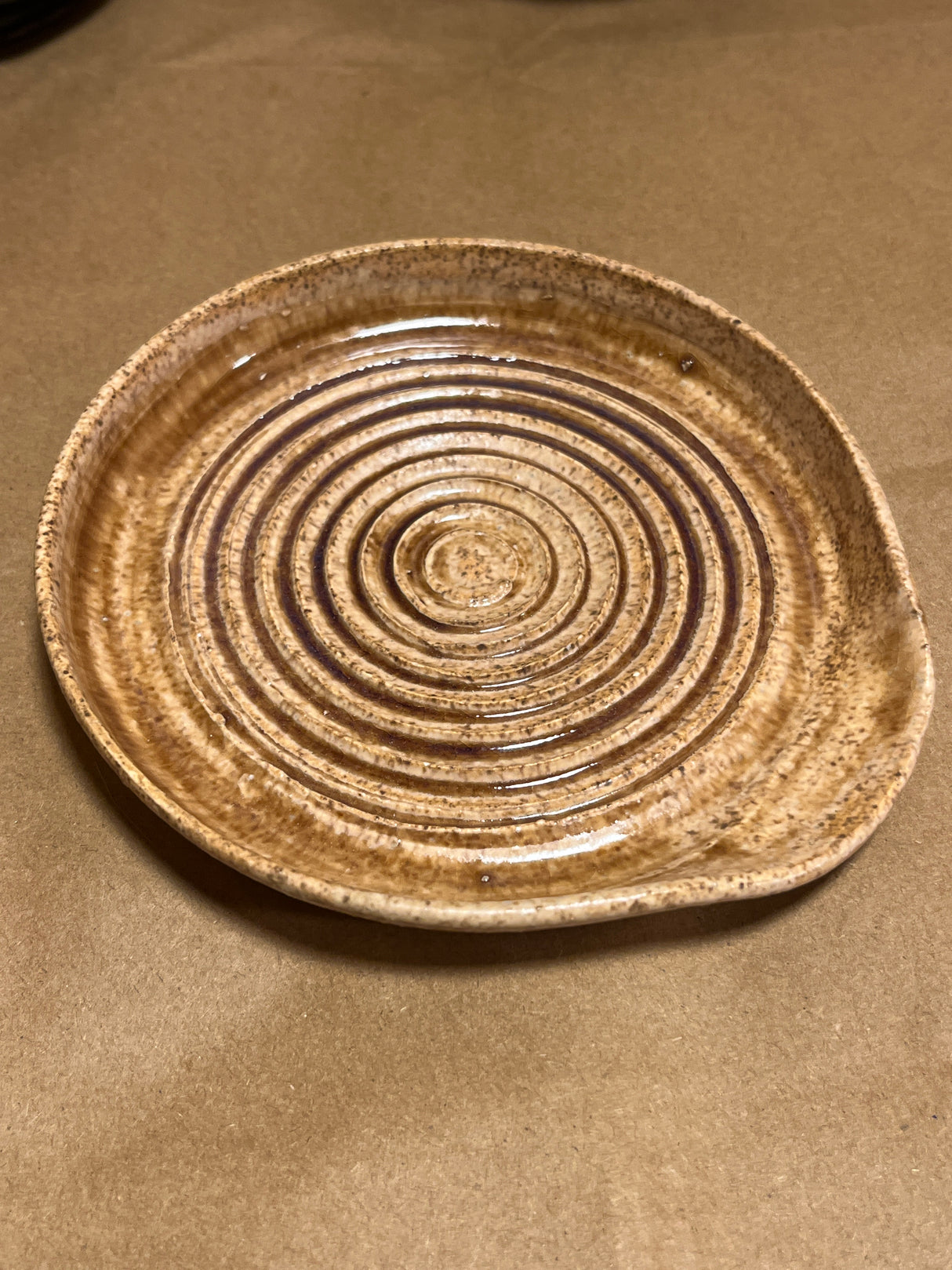 Handmade ceramic garlic grater spoon rest by Charles Lehman, featuring a speckled brown glaze and concentric ridges, displayed on a neutral tabletop.