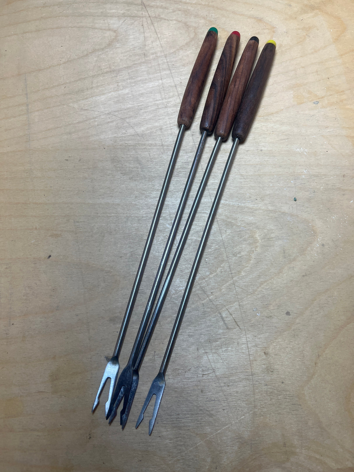 Fondue forks - stainless steel with teak handles and multicolor caps, featuring long metal shafts and two tines, set on a wooden surface.