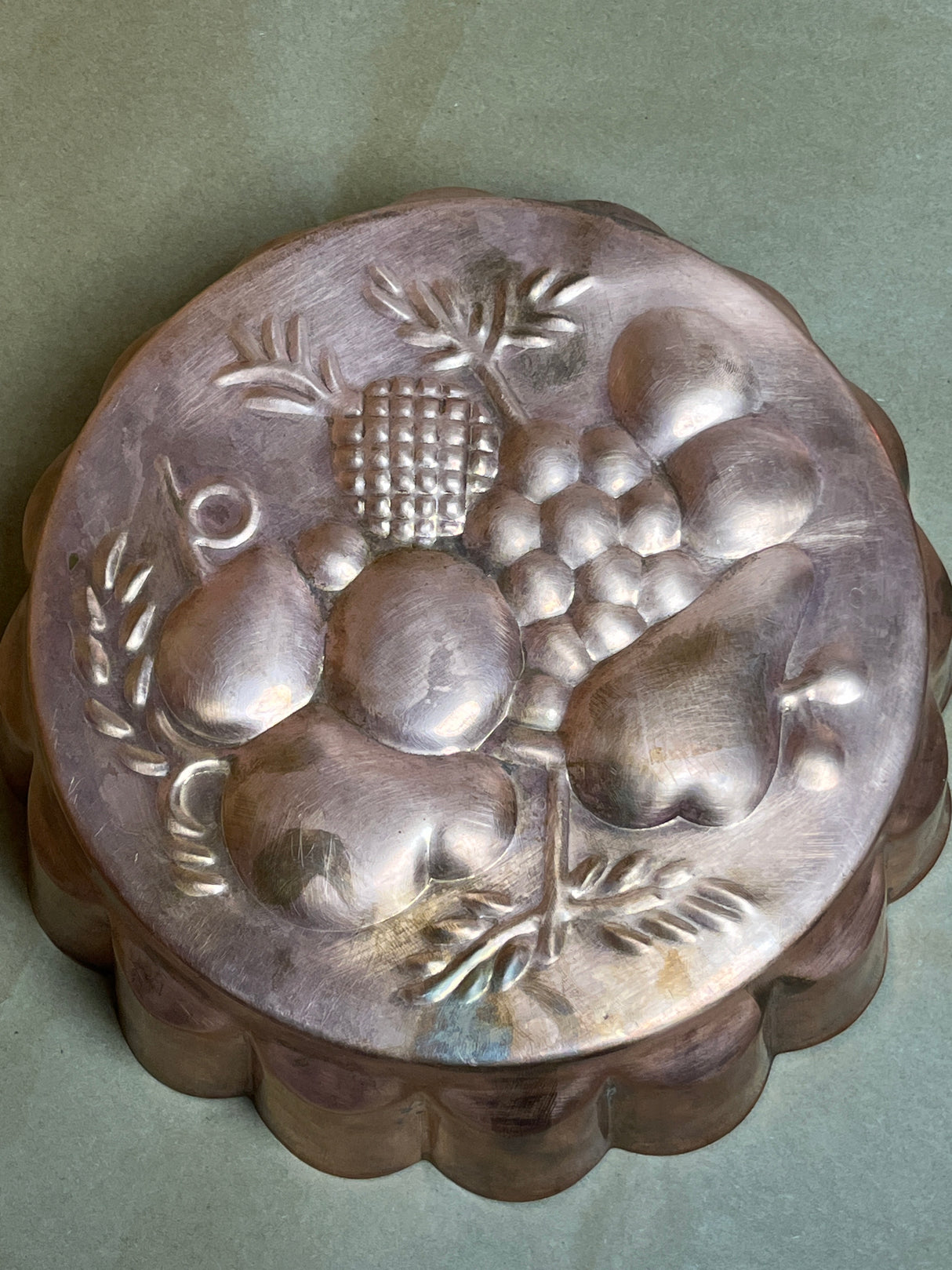 Vintage copper baking pan with embossed fruit design, featuring grapes, pears, and pineapple motifs. Scalloped sides add to its classic charm.