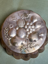 Vintage copper baking pan with embossed fruit design, featuring grapes, pears, and pineapple motifs. Scalloped sides add to its classic charm.
