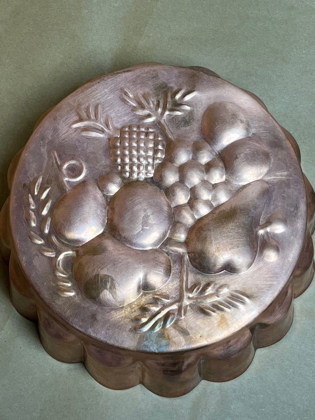 Vintage copper baking pan with embossed fruit design, featuring grapes, pears, and pineapple motifs. Scalloped sides add to its classic charm.