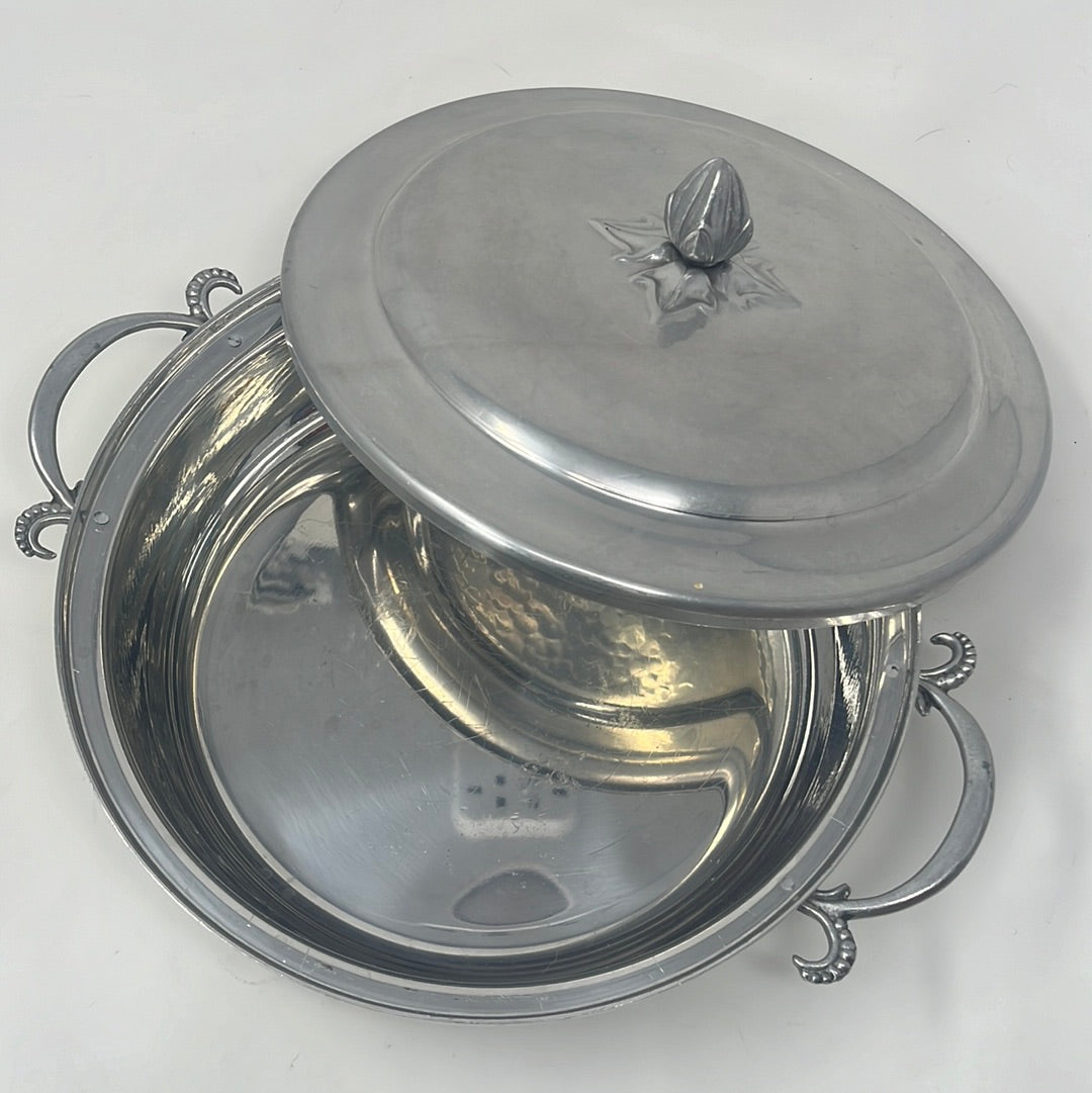 Vintage aluminum casserole carrier with intricate side handles and lid knob, ideal for elegantly presenting baked dishes. Silver bowl with lid on a white surface, featuring metal details.