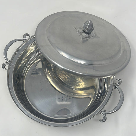 Vintage aluminum casserole carrier with intricate side handles and lid knob, ideal for elegantly presenting baked dishes. Silver bowl with lid on a white surface, featuring metal details.