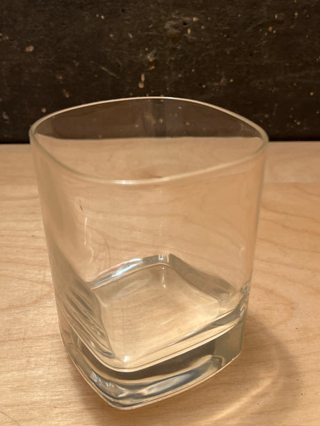 Square heavy bottom tumbler drinking glass with gently rounded square sides, sitting on a light wood surface, perfect for serving drinks with elegance and stability.