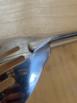 Pedrini stainless steel slotted serving spoon on a wooden surface, showcasing its slotted blade and welded handle, ideal for efficient serving.