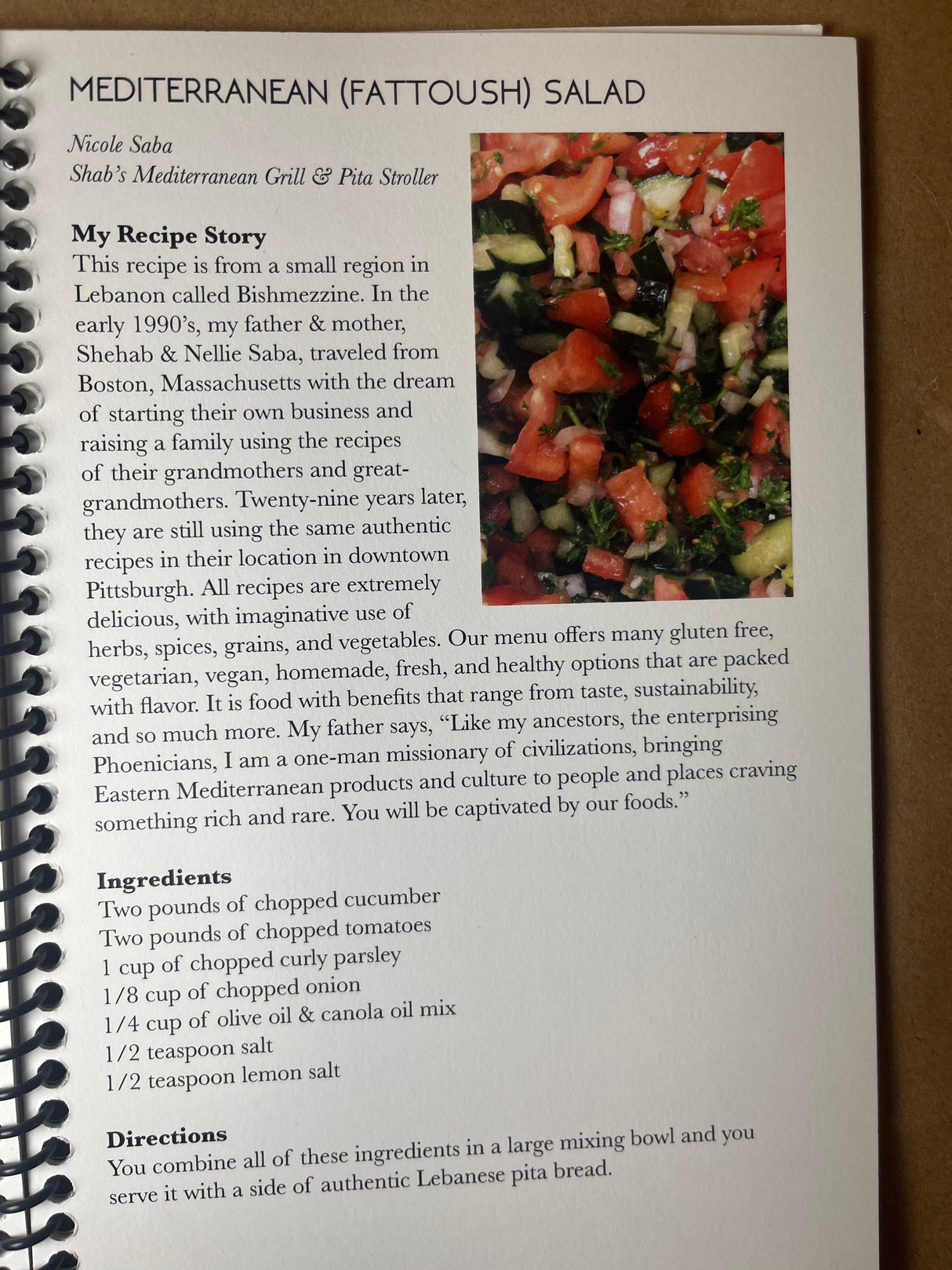 Spiral-bound cookbook open to MEDITERRANEAN (FATTOUSH) SALAD recipe by Nicole Saba, featuring a vibrant salad image and detailed ingredients list from Lebanese heritage.