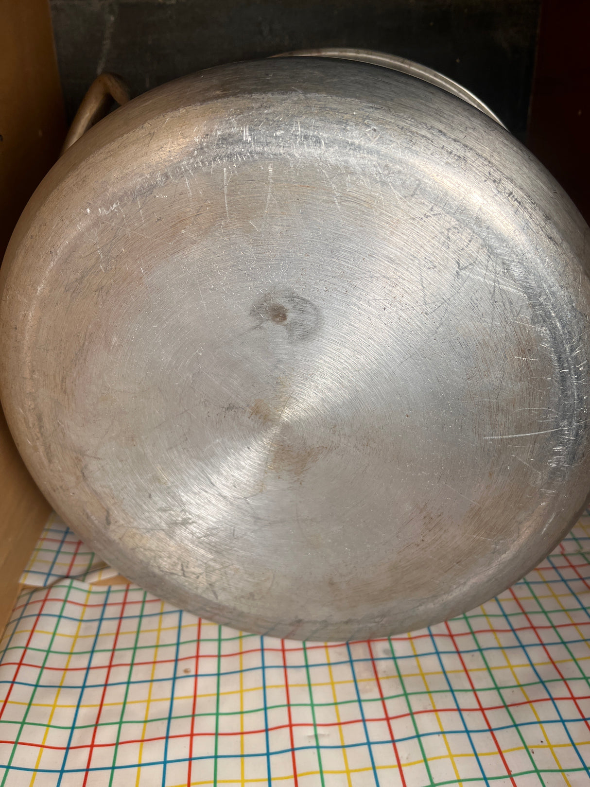 Underside of a vintage Vollrath large stock pot, showcasing its machining marks and surface scratches, resting on a checkered cloth in a storage area.