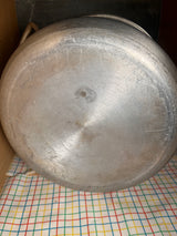 Underside of a vintage Vollrath large stock pot, showcasing its machining marks and surface scratches, resting on a checkered cloth in a storage area.