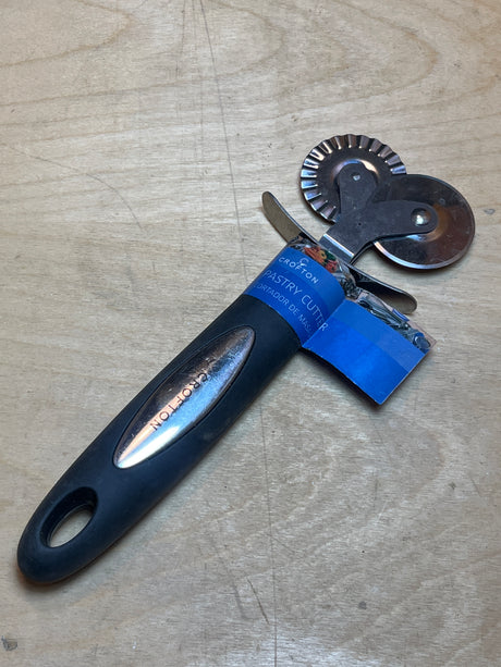 Dual pastry cutter with soft grip black handle from Crofton, featuring straight and fluted wheels, packaged on a wooden surface for slicing and crimping dough.