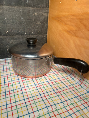 2-qt saucepan with copper bottom - vintage Revere Ware stainless steel, featuring a black plastic handle, sits on a multicolored grid-patterned table covering.