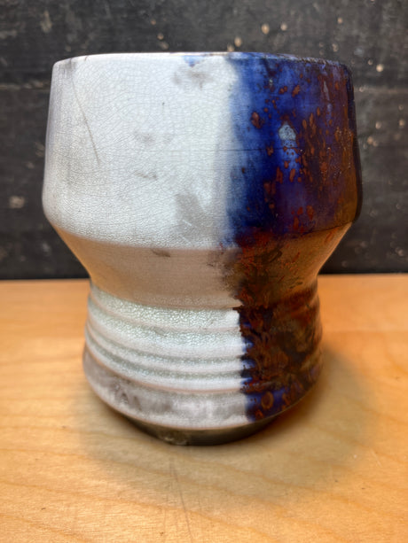 Handmade ceramic utensil holder/vase, featuring a wheel-thrown design with crackled off-white and cobalt-speckled blue-brown glaze, tapering mid-section on a wooden surface.