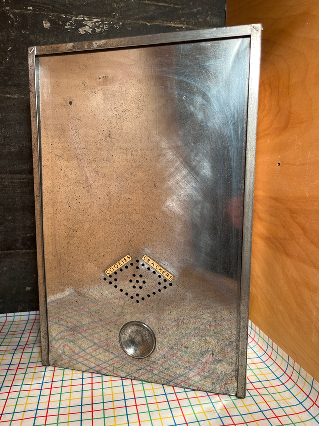 Antique steel bread box drawer insert with sliding lid, featuring ventilation holes, turn-knob, and original COOKIES and CRACKERS labels, set against a patterned tablecloth.