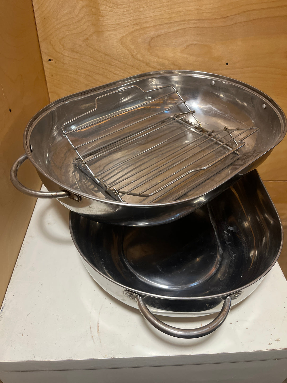 Stainless steel oval roasting pan with rack and lid, featuring two stacked pans with looped handles and a top pan equipped with a removable rack.