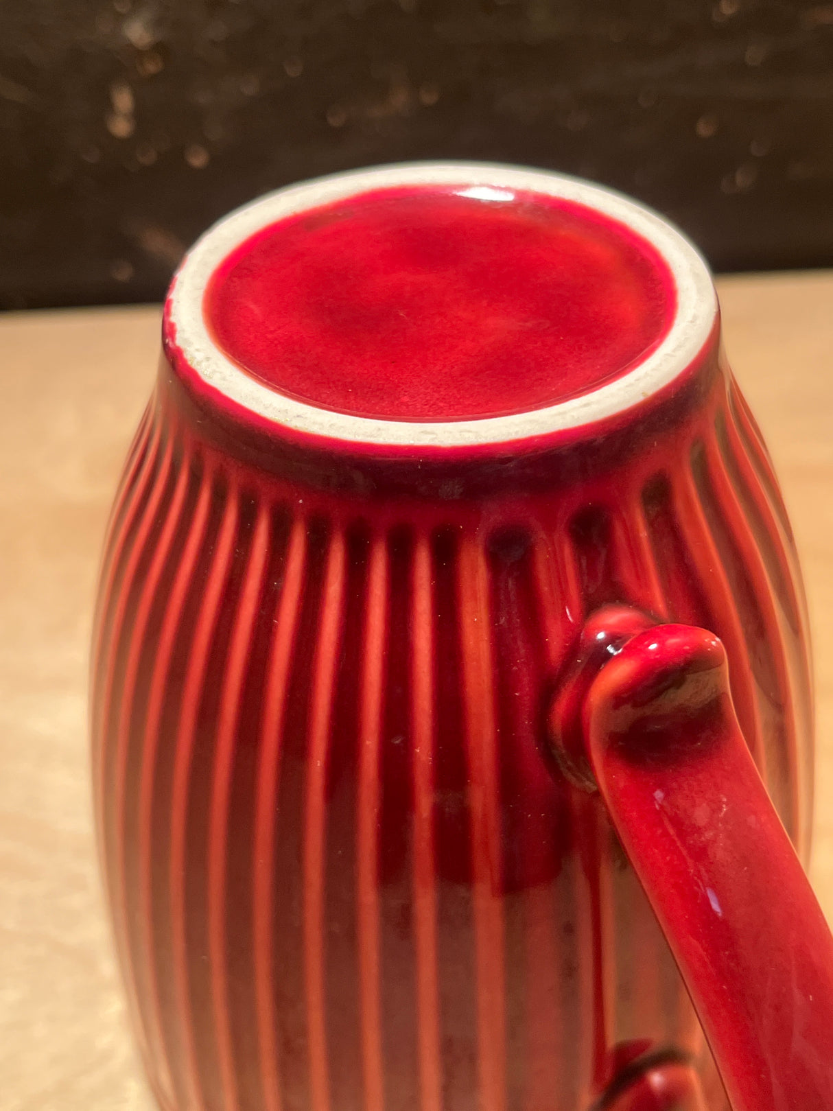 Ribbed ceramic mug, red glazed exterior with black interior, shown upside-down on a wooden surface, highlighting its circular unglazed base and side handle.