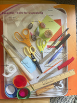 What’s New, Cupcake? cookbook surrounded by various crafting tools, including scissors and a ruler, highlighting creative baking ideas and recipes.