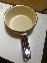 Tan and brown enameled sauce pot, featuring a retro design with a chip on the handle, and a notch for easy hanging storage.