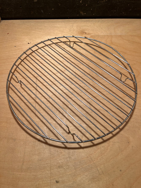Circular wire rack for cooling, roasting, or campfire cooking, displayed on a wooden work surface, showcasing its sturdy stainless-steel construction and versatile design.