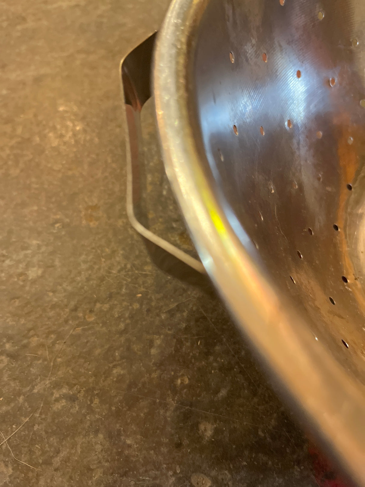 Close-up of a Colander with flat base, showcasing its perforated stainless steel body and one handle on a countertop. Note the lightweight design and shallow base.