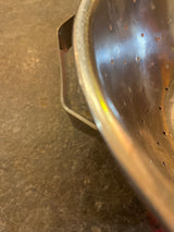 Close-up of a Colander with flat base, showcasing its perforated stainless steel body and one handle on a countertop. Note the lightweight design and shallow base.