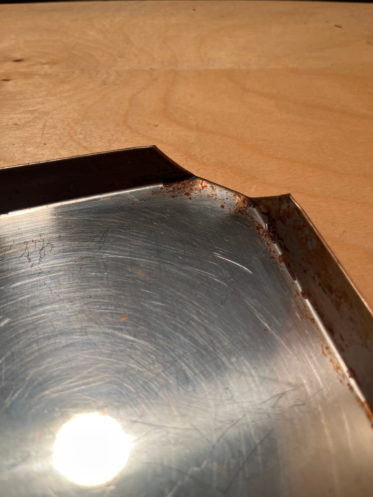 Close-up of a vintage Sears stainless steel baking pan with circular scratches and light rust on edges, reflecting overhead light on its surface.