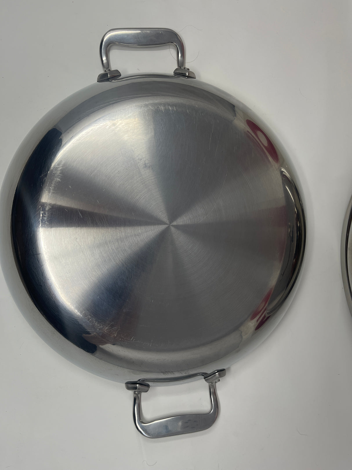 All-Clad stainless steel paella braise pan, featuring a close-up of its double handles and metal surface, ideal for cooking with a domed lid.