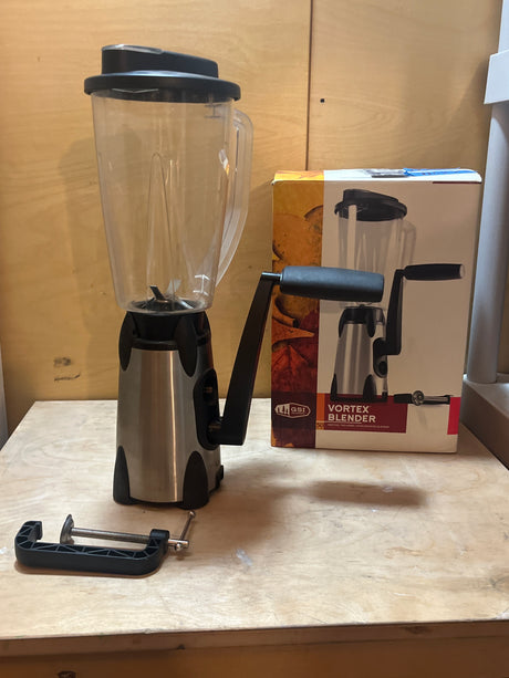 GSI Outdoors Vortex hand-cranked blender with clear jar, black lid, stainless-steel base, and hand crank. Includes detachable C-clamp and original retail box in background.