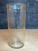 Anchor Hocking narrow glass jelly jar, a tall, transparent tumbler, sits empty on a wood surface, showcasing its sleek cylindrical design against a textured backdrop.