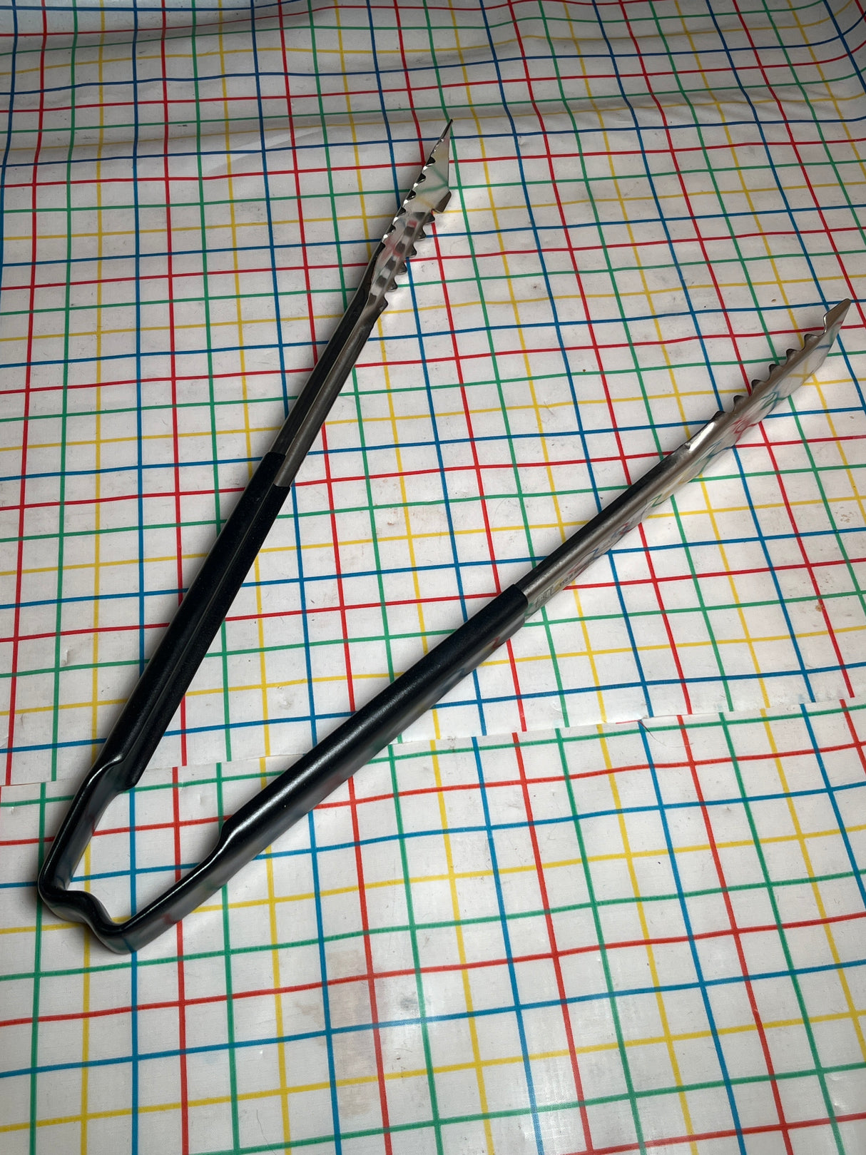 Two stainless-steel serving tongs with black Kool-Touch® handles, 16 inches long, lay on a multicolored grid-patterned surface; VersaGrip Vollrath Jacob's Pride® model.