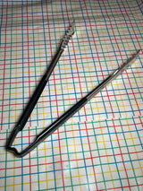 Two stainless-steel serving tongs with black Kool-Touch® handles, 16 inches long, lay on a multicolored grid-patterned surface; VersaGrip Vollrath Jacob's Pride® model.