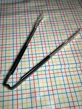 Two stainless-steel serving tongs with black Kool-Touch® handles, 16 inches long, lay on a multicolored grid-patterned surface; VersaGrip Vollrath Jacob's Pride® model.