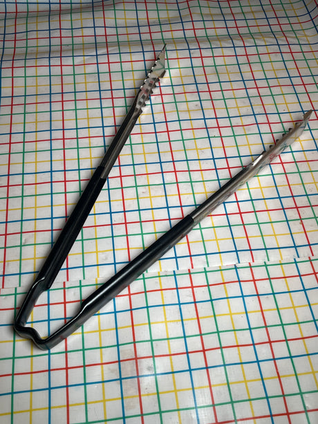 Two stainless-steel serving tongs with black Kool-Touch® handles, 16 inches long, lay on a multicolored grid-patterned surface; VersaGrip Vollrath Jacob's Pride® model.