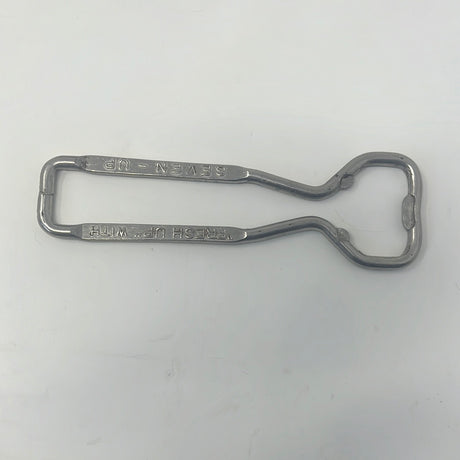 Vintage bottle opener with Fresh Up with Seven-Up inscription, alongside tongs and a metal sign on a white surface.