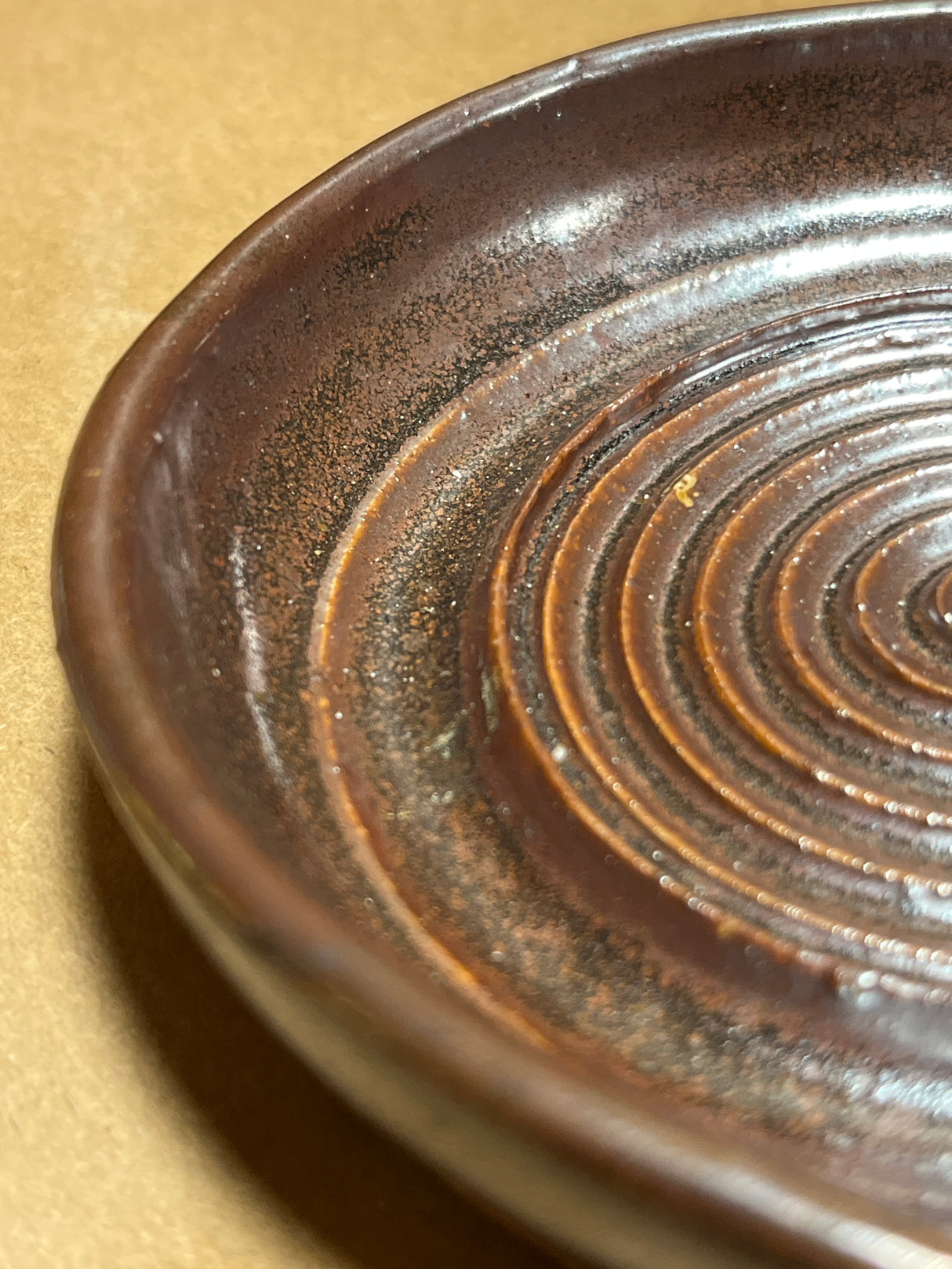 Handmade ceramic garlic grater spoon rest by Charles Lehman, featuring a brown, speckled glaze with concentric ridges, on a kraft-paper-like surface.