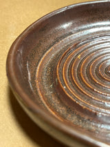 Handmade ceramic garlic grater spoon rest by Charles Lehman, featuring a brown, speckled glaze with concentric ridges, on a kraft-paper-like surface.