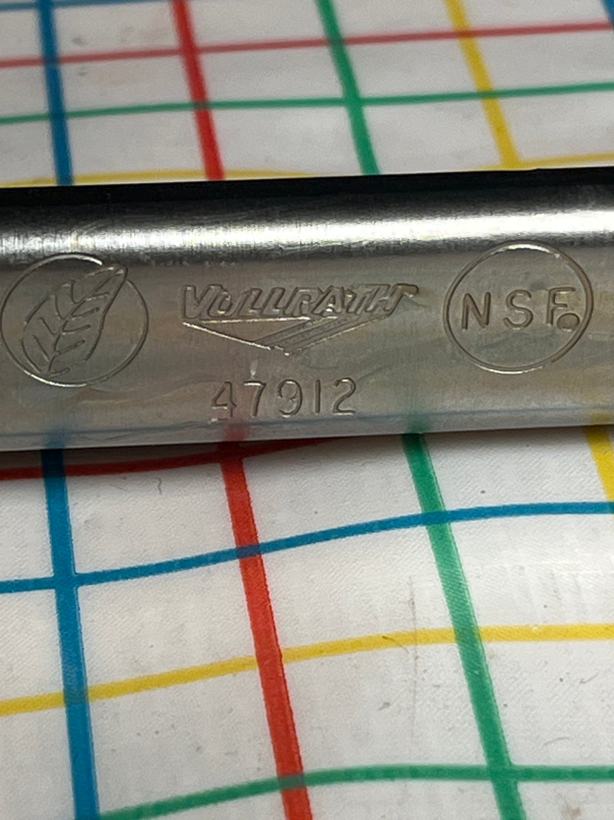 Close-up of Vollrath 4791620 stainless steel serving tongs handle, featuring NSF mark and leaf symbol, resting on patterned cloth.