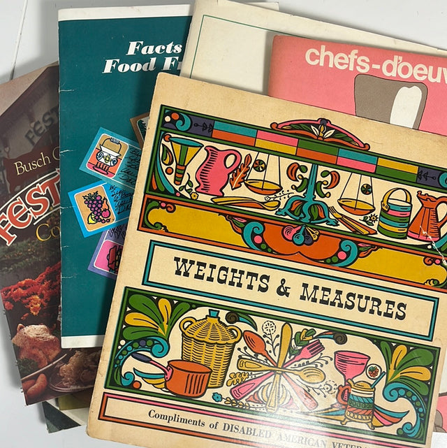 Mystery cookbooklet on a table, featuring colorful graphics and surrounded by various books and cards, inviting culinary exploration for just one dollar.