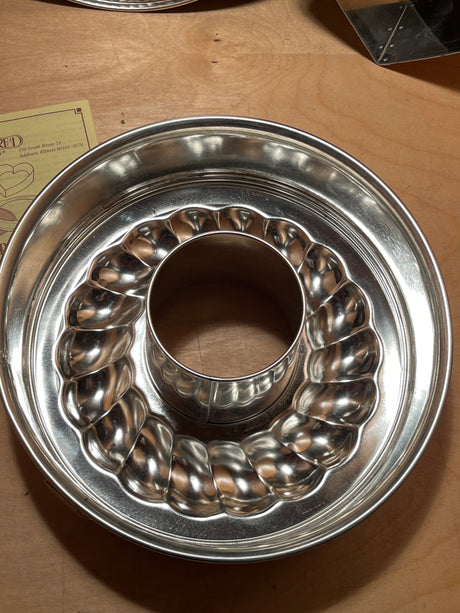 4-piece springform pan set by Pampered Chef, featuring a shiny, fluted Bundt-style cake pan on a wooden surface, partially covering a small paper flyer.