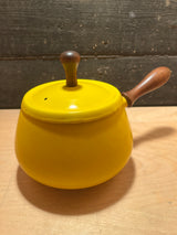 Vintage enamel fondue pot in sunshine yellow with wood handles, featuring a wooden knob lid, rests on a light wood surface against a textured backdrop.