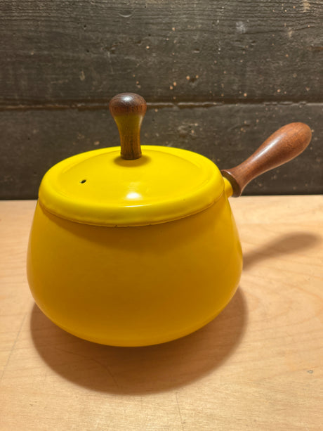 Vintage enamel fondue pot in sunshine yellow with wood handles, featuring a wooden knob lid, rests on a light wood surface against a textured backdrop.