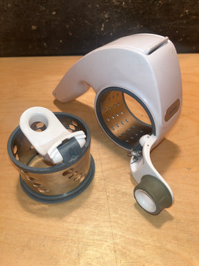 Rotary grater with two drums - Zyliss, featuring a white plastic housing with a crank handle and two interchangeable stainless-steel grating drums, disassembled for display.