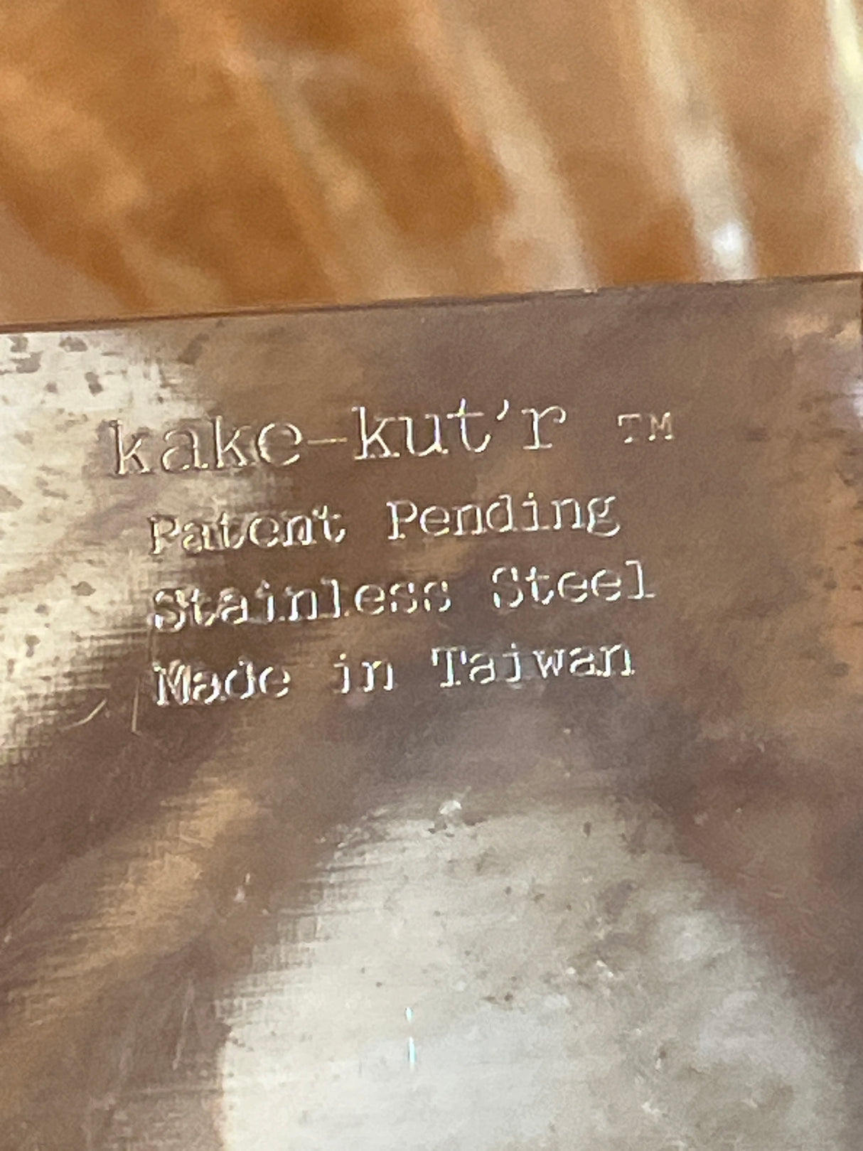 Square Kake-kut’r close-up showing metal stamp details: “kake-kut’r™,” “Patent Pending,” “Stainless Steel,” “Made in Taiwan.” Ideal for easy one-handed dessert serving.